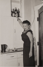 Black and white photo of a woman grabbing a bottle of liquor from a kitchen cabinet.