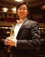 Musician Apollo Lee poses in a tuxedo in a concert hall, holding his trumpet to his chest. He is wearing glasses and slightly smiling.